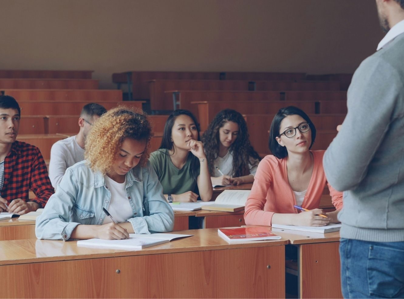 A group of students sitting at desks in a classroom, listening to a teacher.