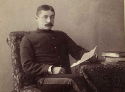 A man in a vintage military uniform sits on a chair, holding a letter with books on a table.