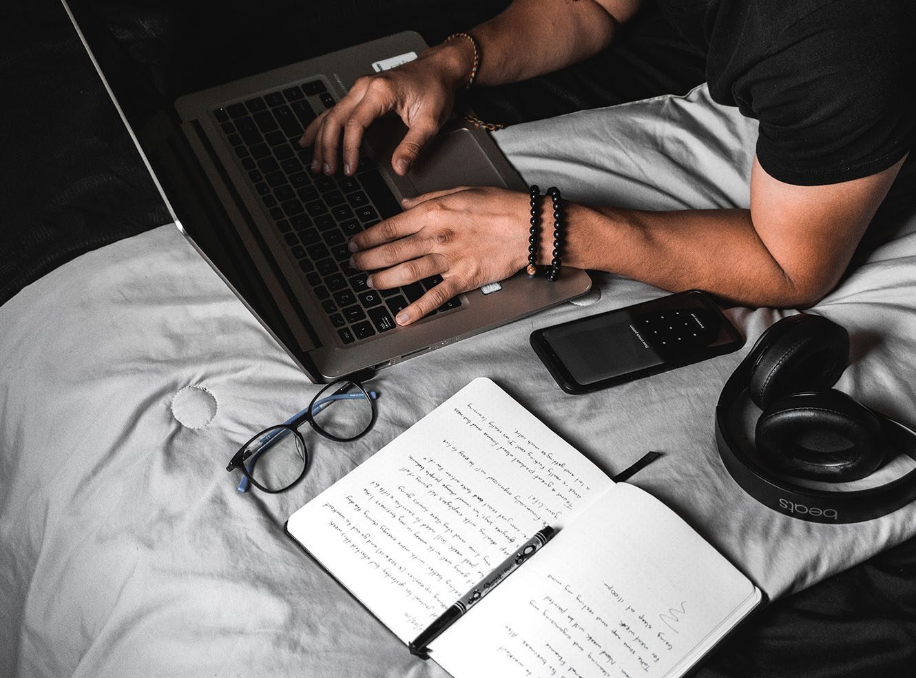 A person lying in bed with a laptop, notebook, headphones, glasses and smartphone.