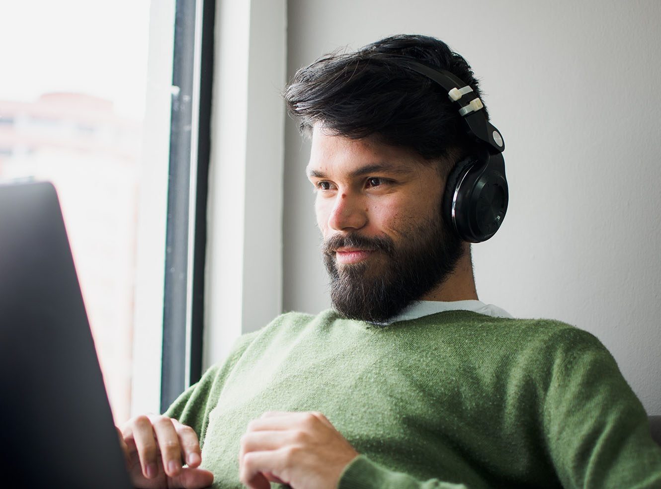 A man with headphones on sitting by a window, focusing on a laptop.