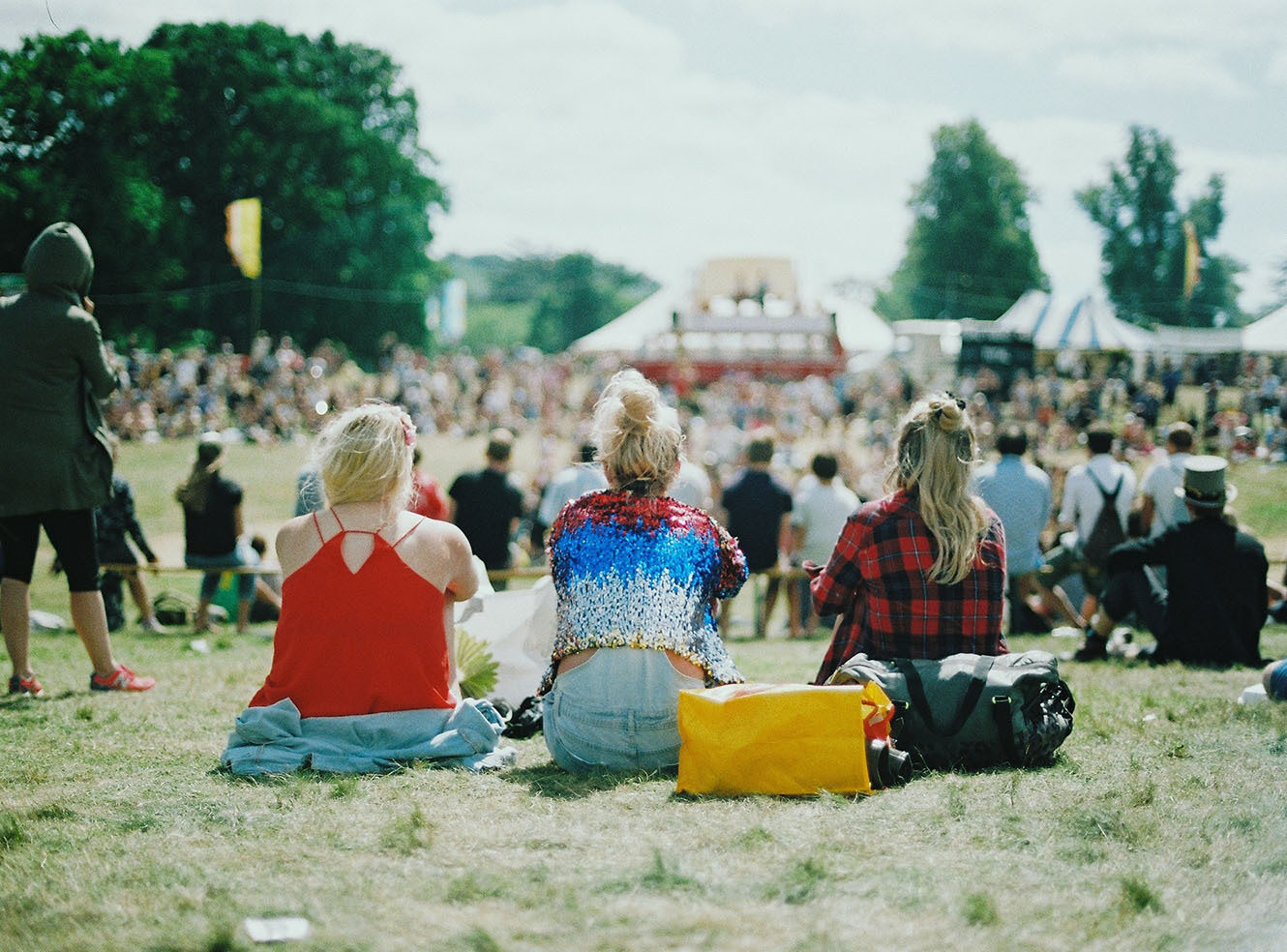 Three people sit on the grass at an outdoor festival, facing a large crowd with tents in the background. A yellow bag lies beside them