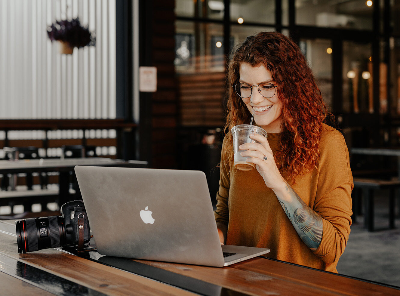 A woman with curly hair and glasses enjoys a drink while working on a laptop in a cafe