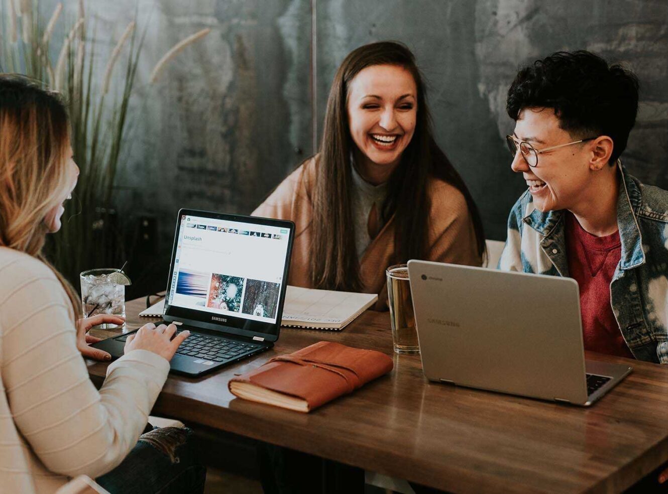 Three friends studying at a wooden table with their laptops, laughing and enjoying drinks.