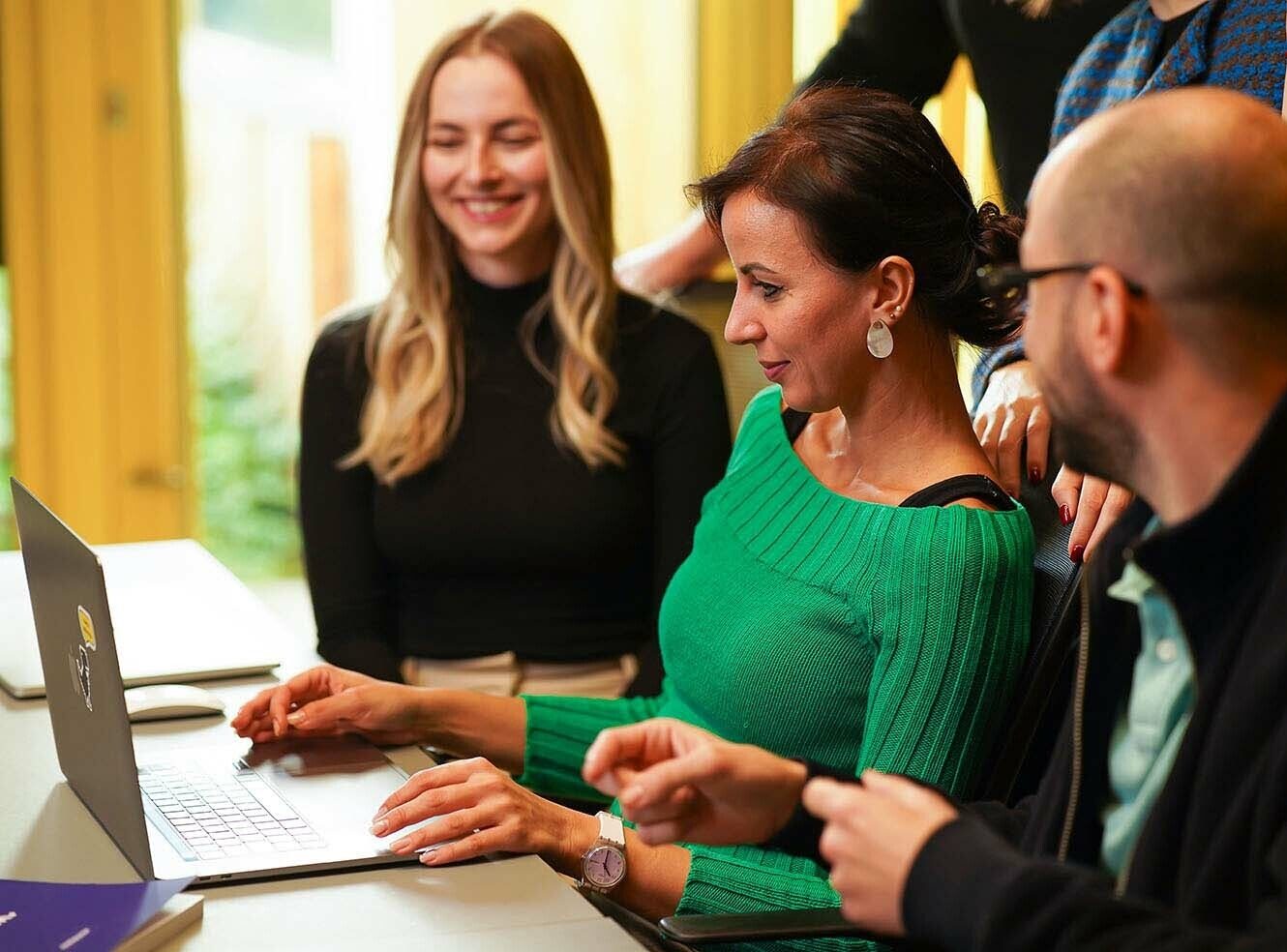 Three people collaborating around a laptop, with one woman in a green sweater actively working.