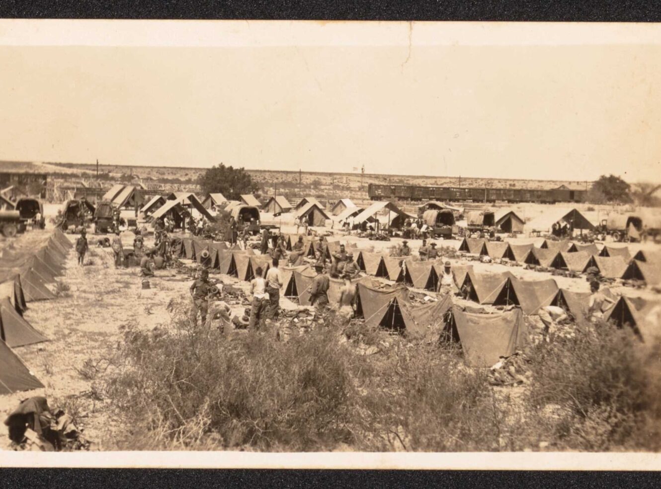 A large outdoor military camp with rows of tents and people working. Trucks are parked nearby.