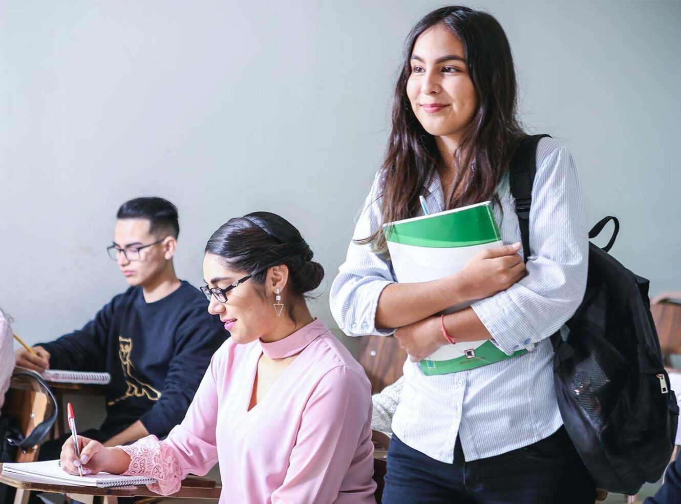 A group of students in a classroom. One girl holds a notebook and smiles while standing, others are writing.