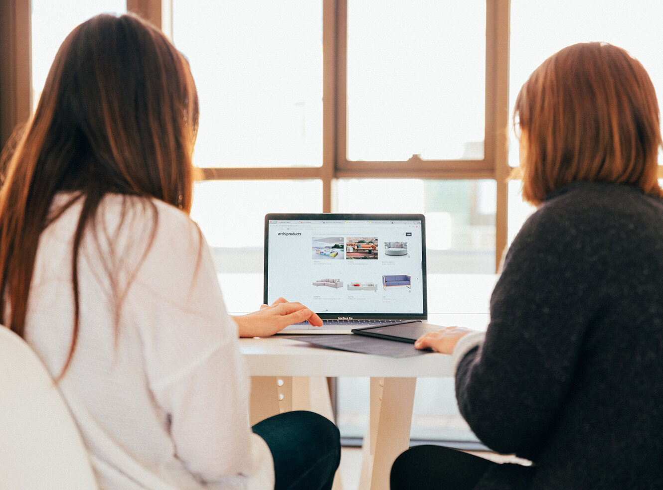 Two women sitting at a table, working on a laptop with bright window light.