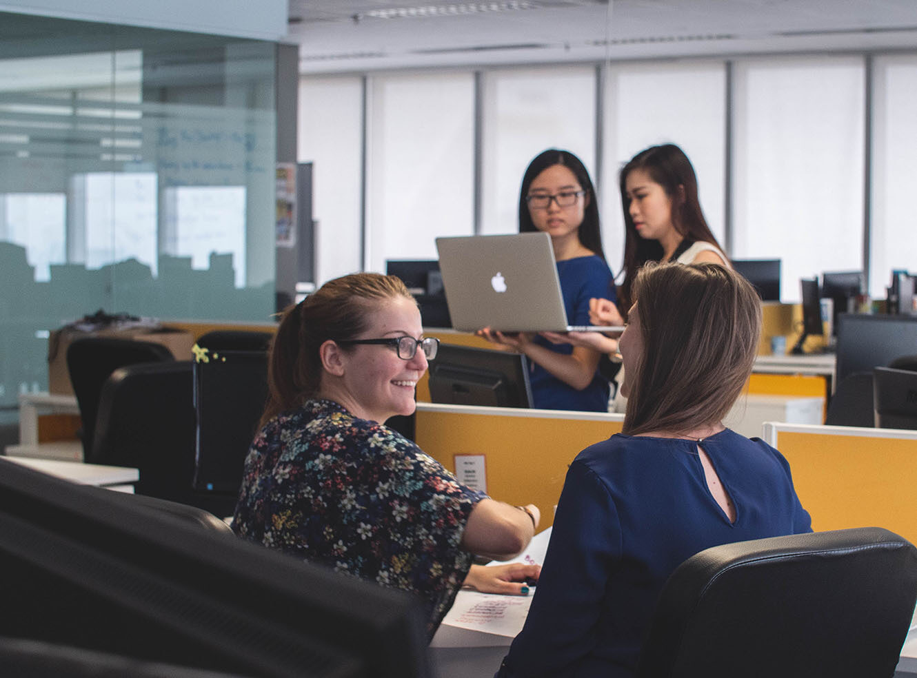 An office with two women seated at desks in the foreground, engaged in discussion, and two standing in the background reviewing a laptop