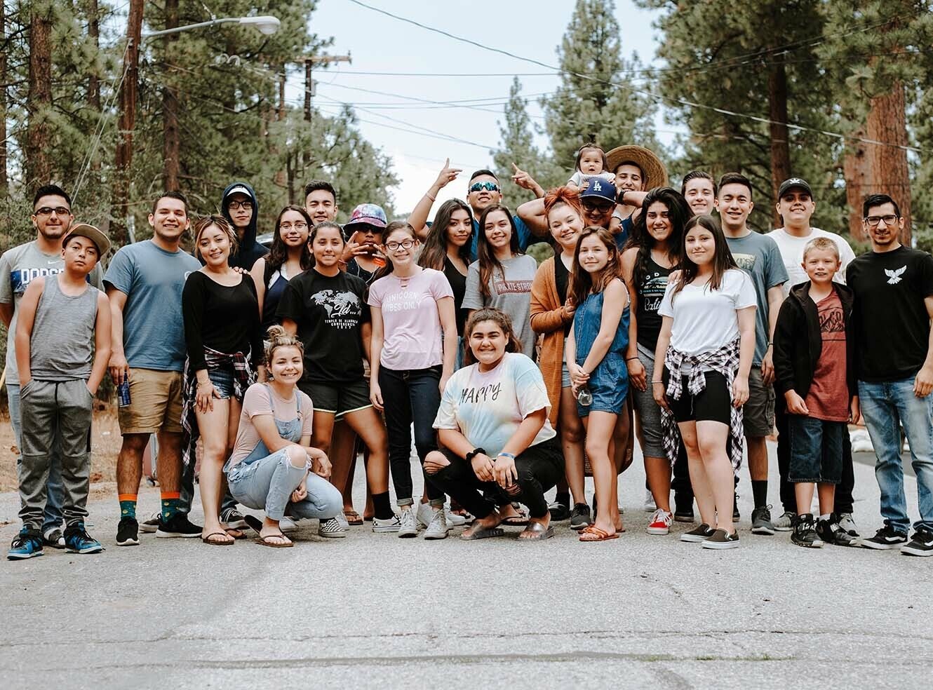 A large group of young people and adults posing together on a road surrounded by tall trees