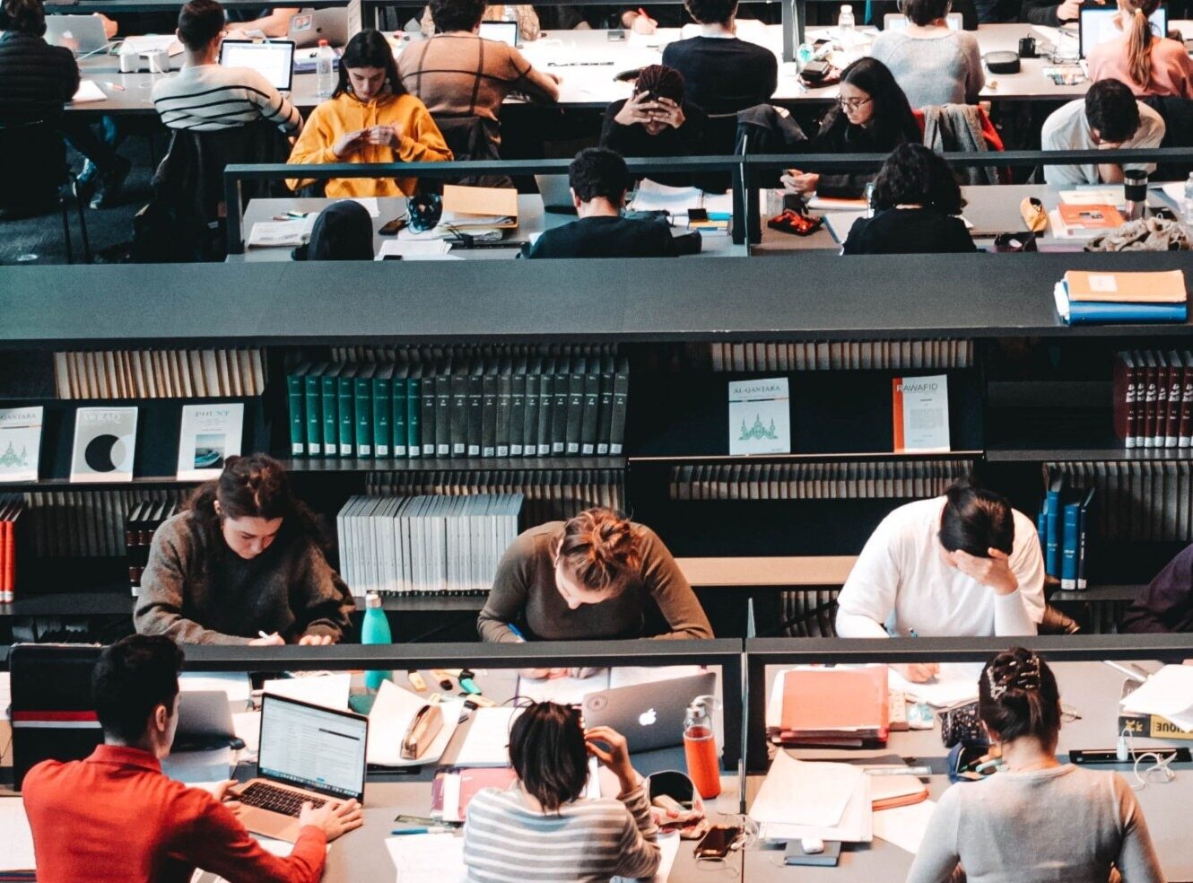 Students work at desks and laptops in a university library study area, with bookshelves and academic materials visible in the background.