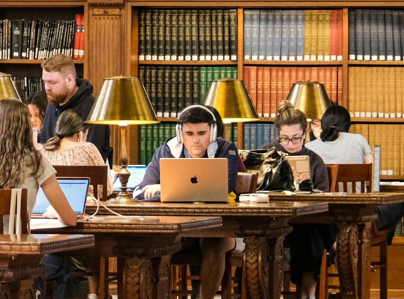 Students studying in a library with bookshelves and desk lamps.