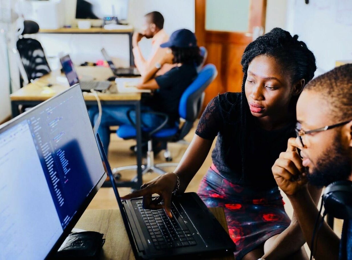 A woman points to a laptop screen while discussing with a man. Other people work in the background