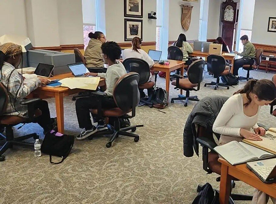Students studying at desks in a library, using laptops and writing notes.