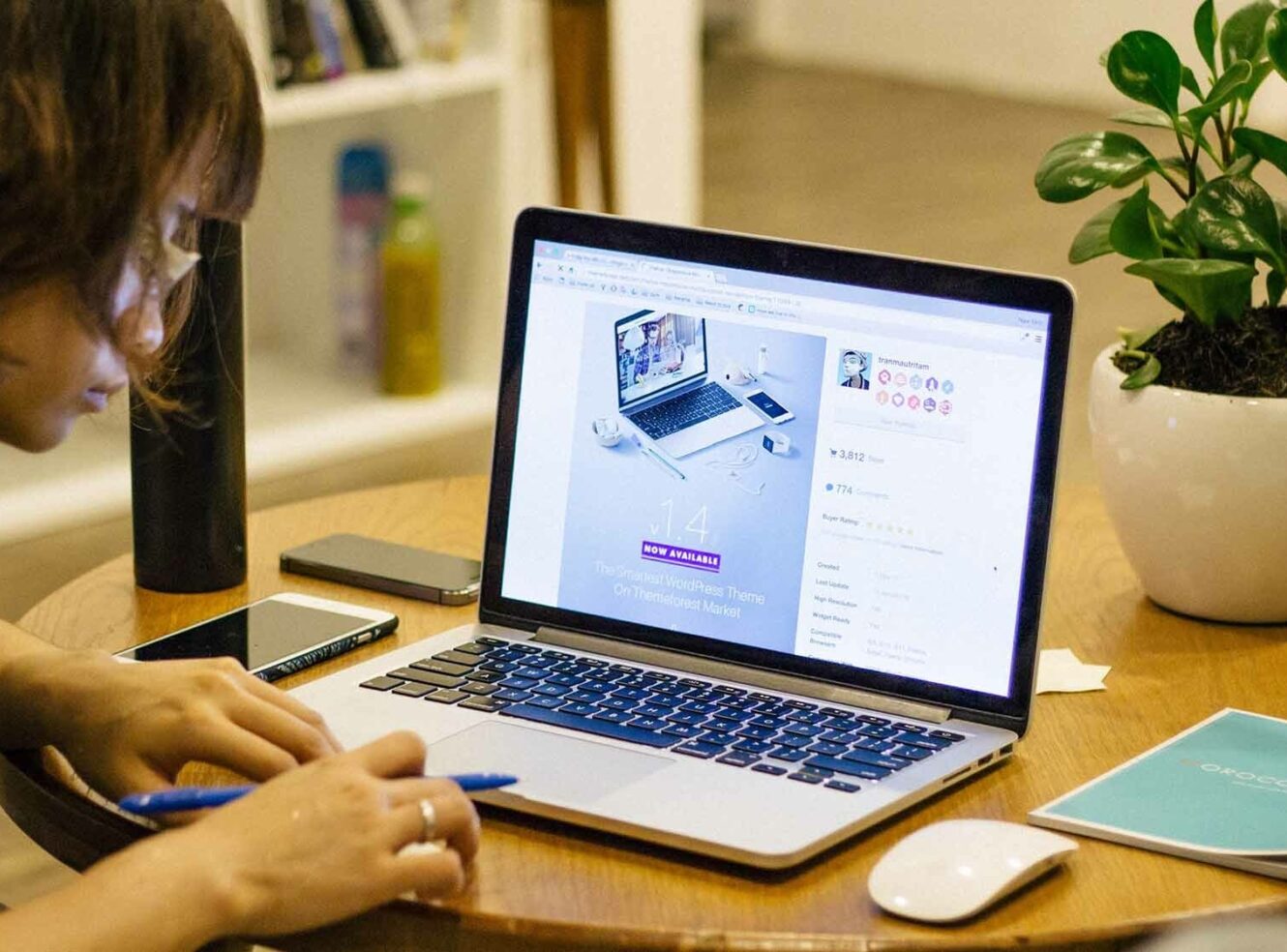 A person using a laptop on a wooden table with a pen in hand. A smartphone and plant nearby.