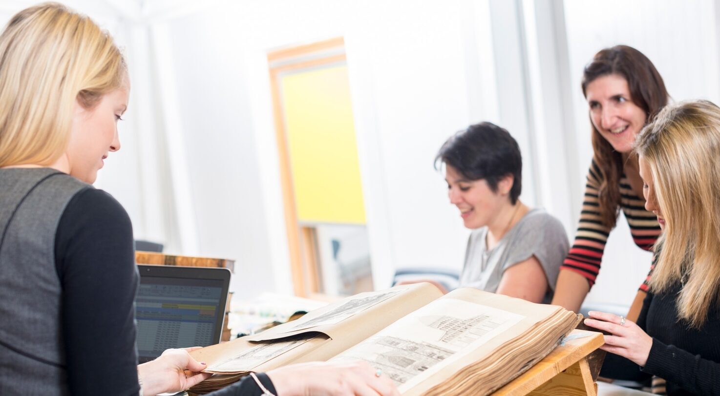 A group of four women are gathered around a large book, discussing the pages. One woman holds the book open, showing illustrations.