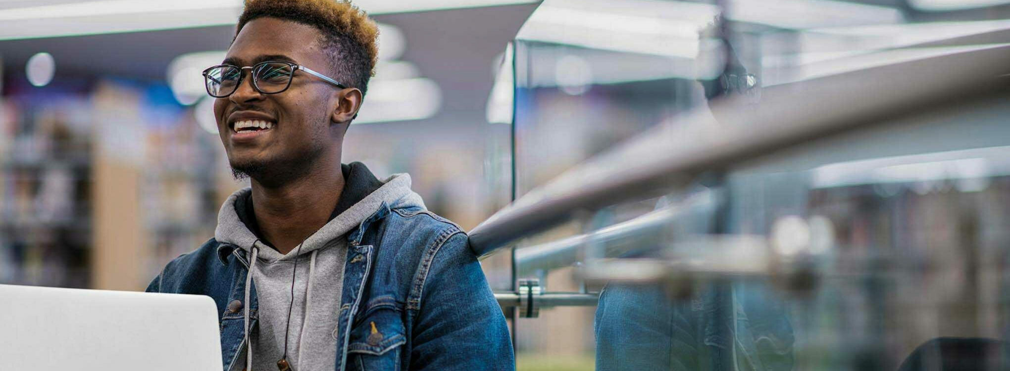 A smiling young man with glasses working on a laptop in a library.