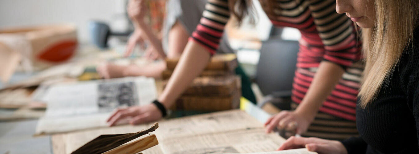 Two people examining old books and documents on a table in a workspace.