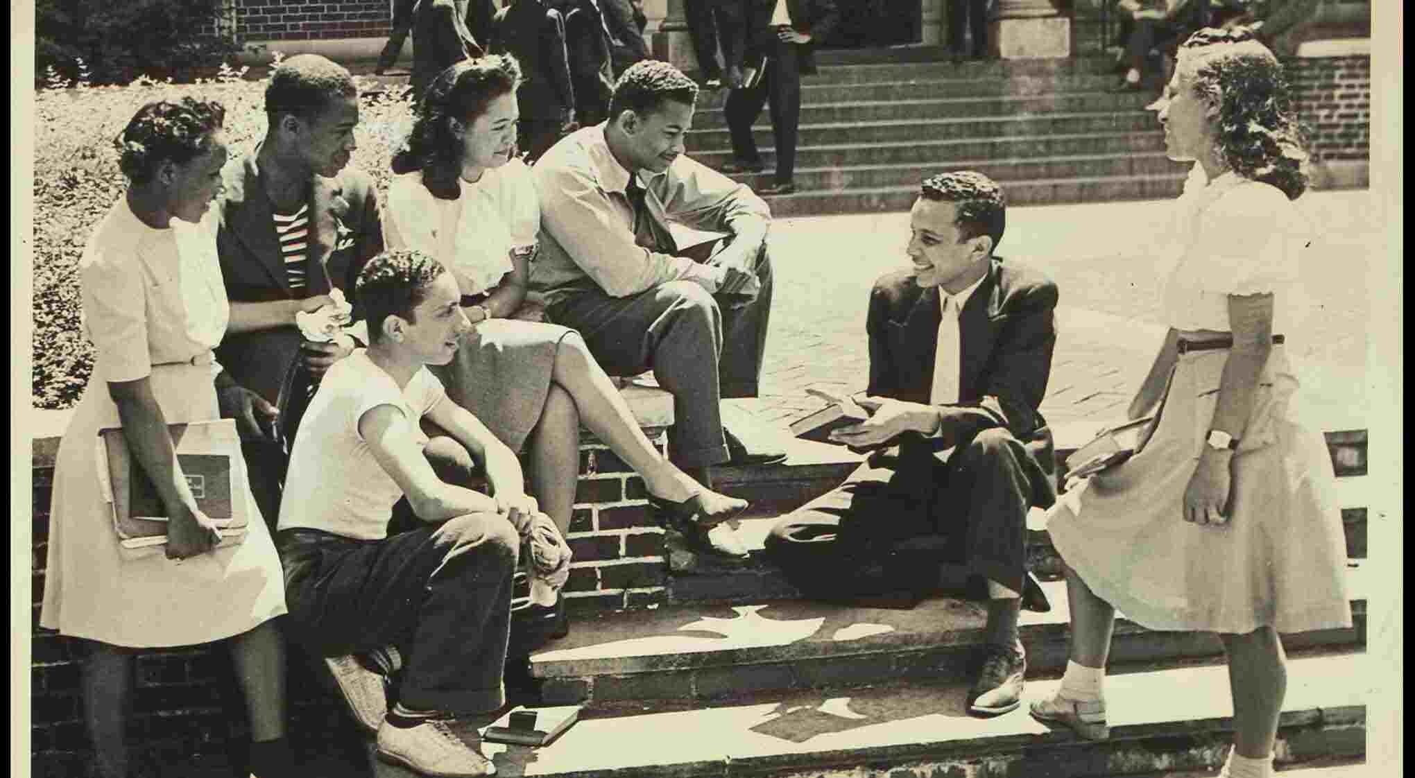 A group of six Black students sitting on steps, engaged in conversation and reading books.