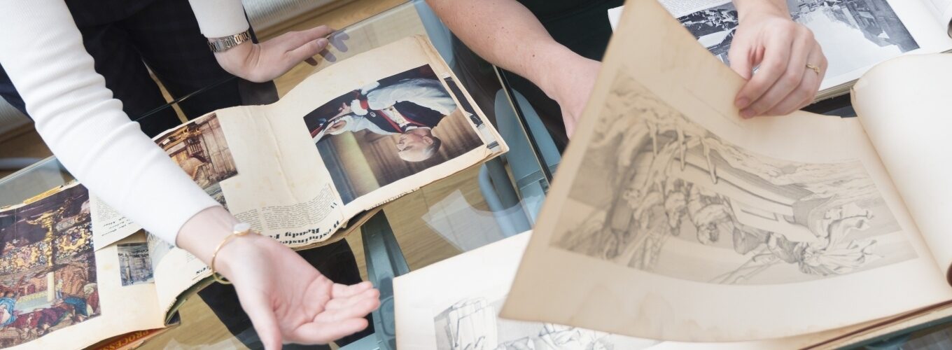Two people examining old books and artwork on a glass table with various pages visible.