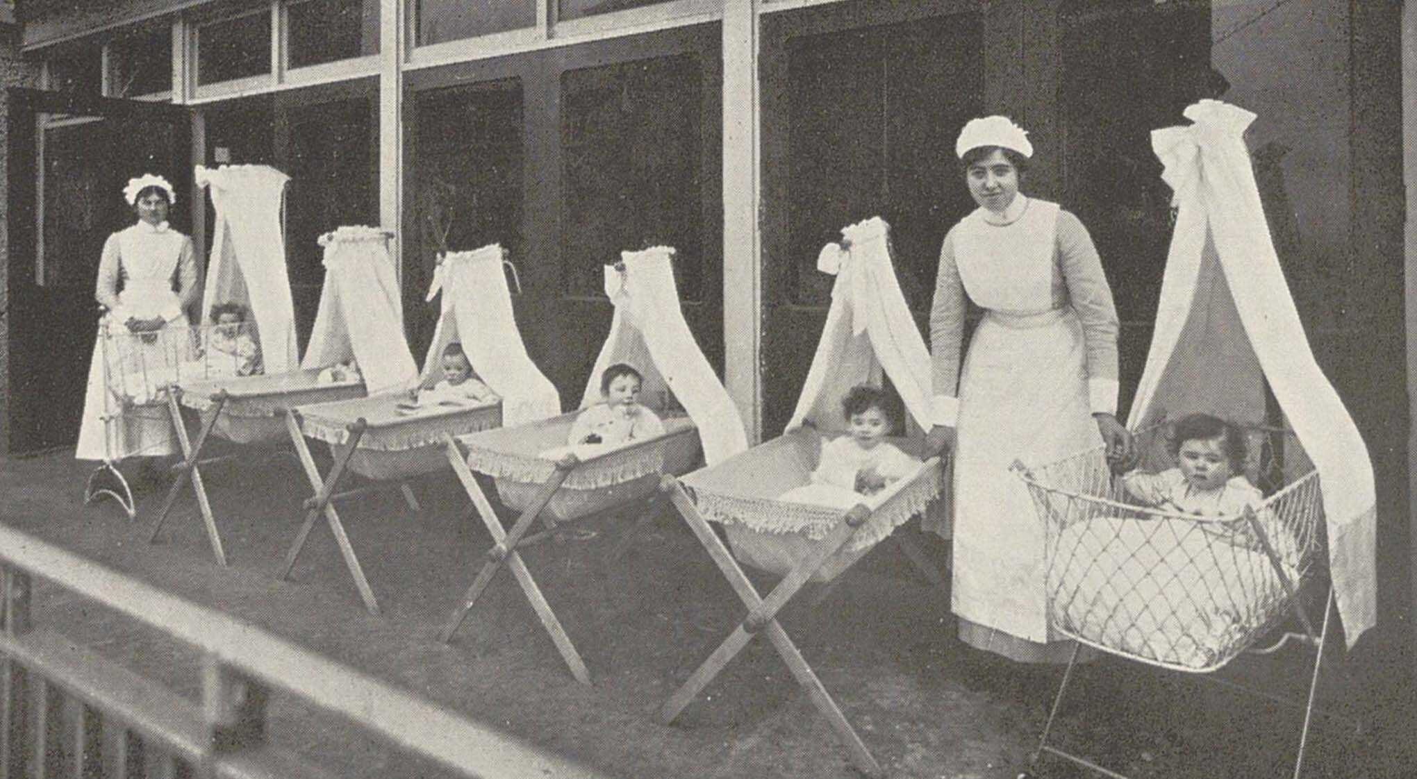 Two women in white nurse uniforms stand by cradles with infants cradles, outdoors.