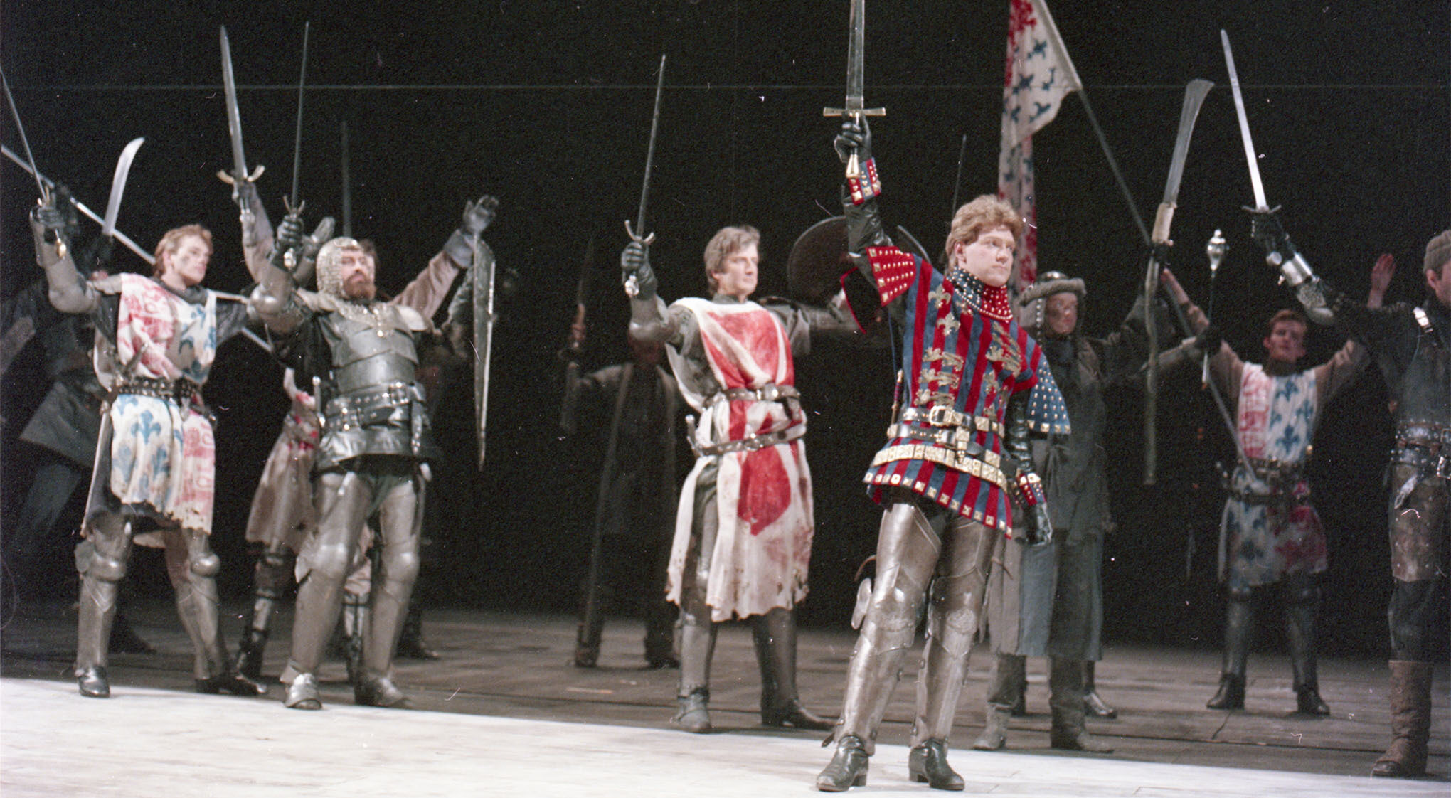 Actors in medieval armour raising swords on stage, showcasing a dramatic scene from Henry V.