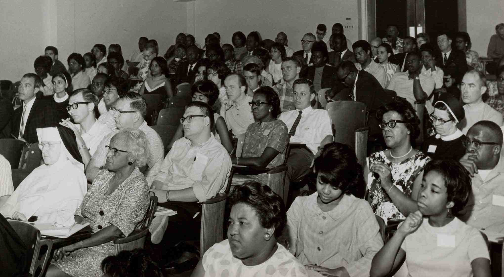 A greyscale photograph of a diverse group of people seated in an auditorium, focused on a speaker.