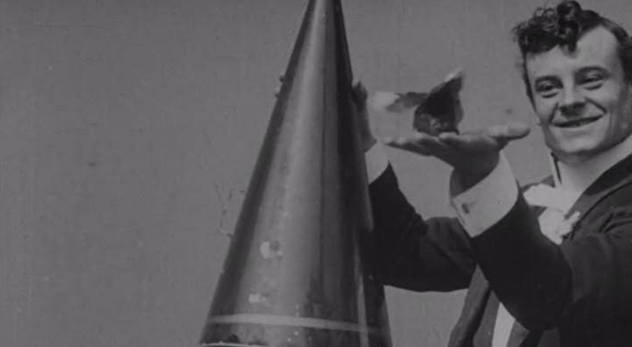 A magician in a suit holding a large hat in one hand, and a bird in the other.