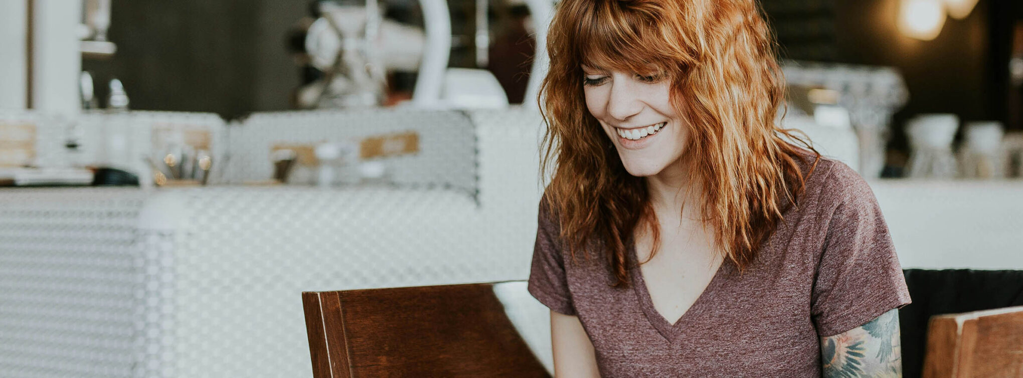 A smiling woman with red curly hair working on a laptop in a cozy café.