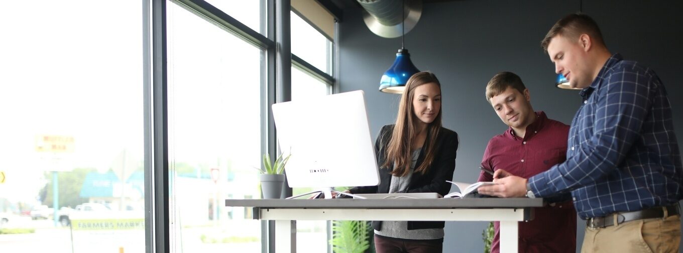 Three people discussing at a standing desk, with a computer and a notebook.